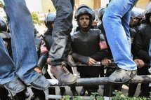 Egyptian protesters stand on a security fence as riot police watch.